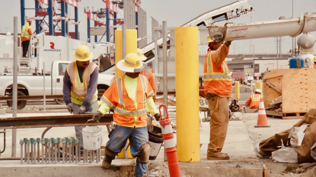 A construction crew placing concrete alongside rail track at a port terminal, gantry cranes behind them